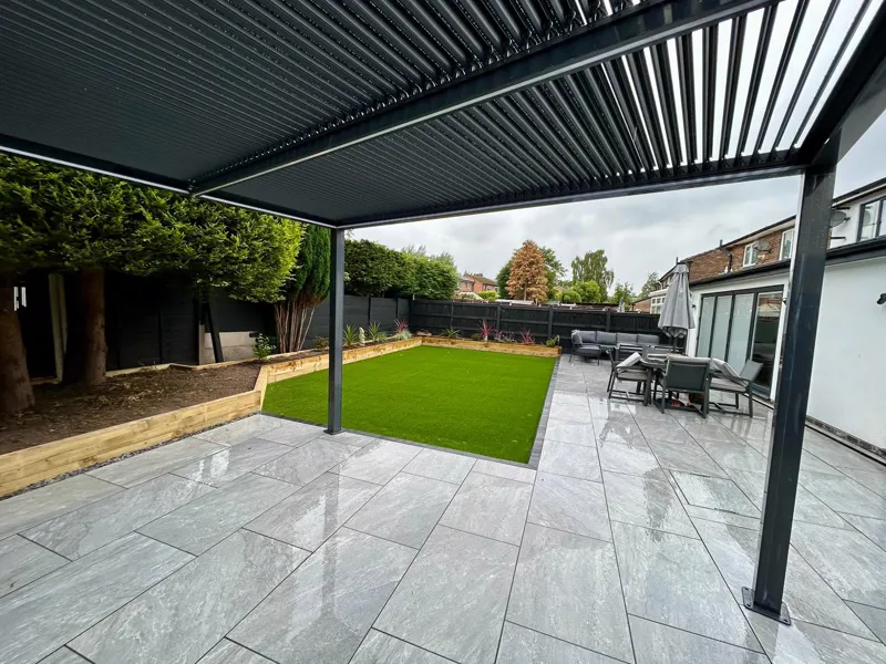 View from beneath a louvred pergola across wet porcelain paving and lawn